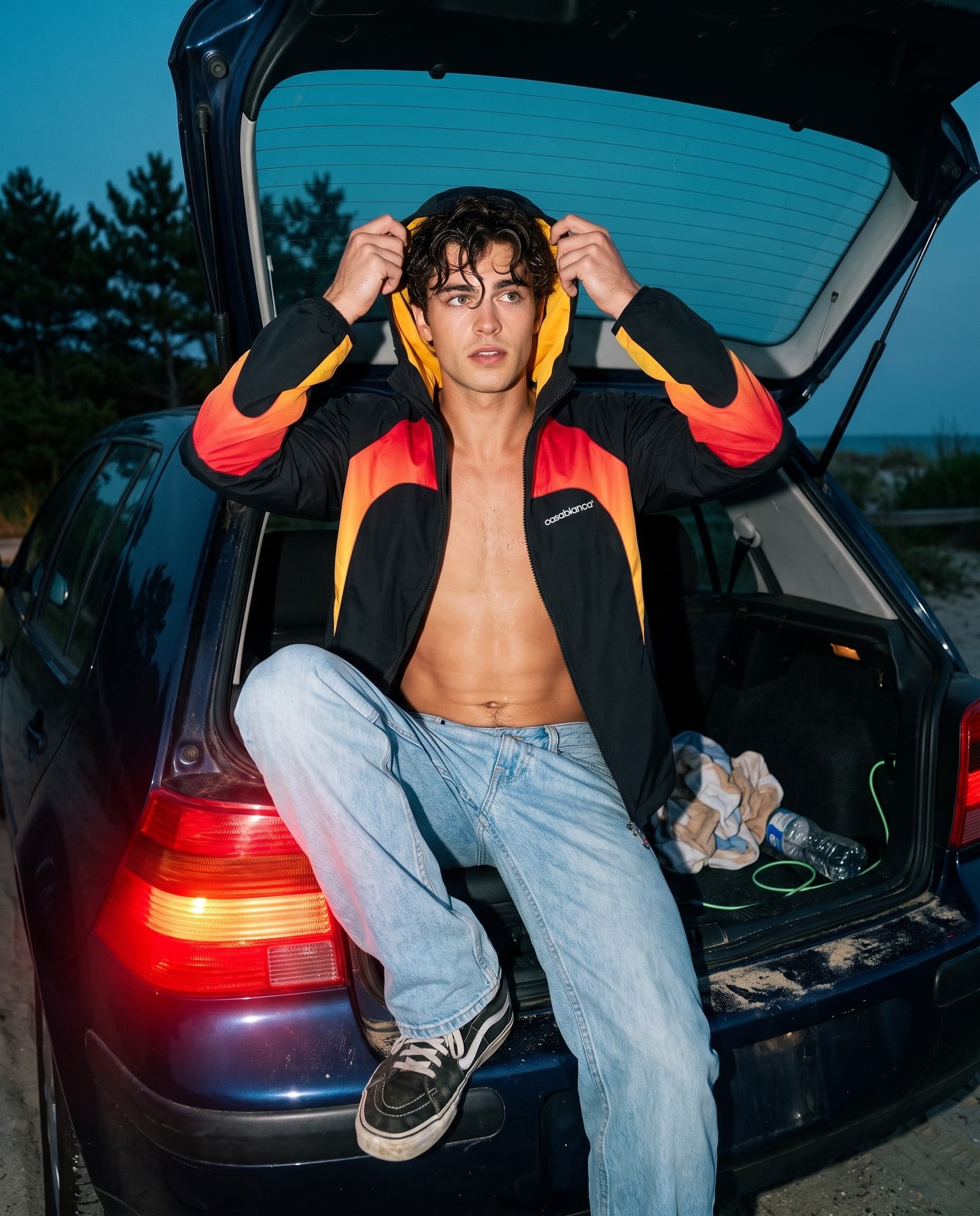 Young man in a flame-panel jacket pulling up the hood while sitting in an open hatchback trunk, lit by flash at dusk, made with Nano Banana Pro