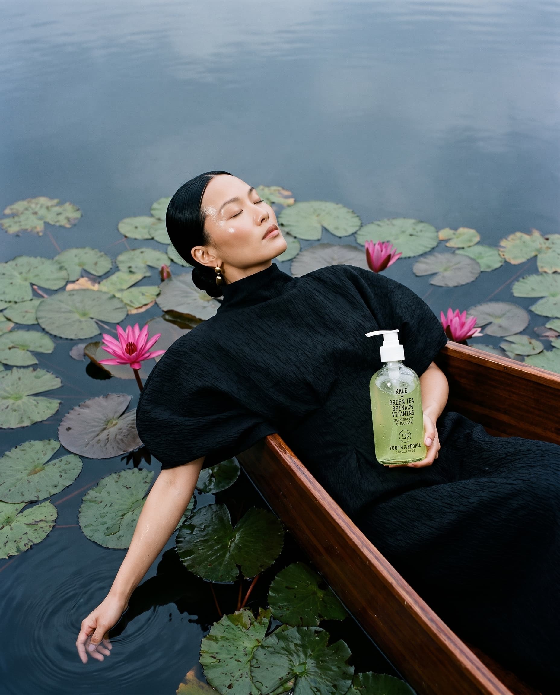 Woman in a black sculptural dress reclining in a wooden boat among pink lotuses, a green cleanser bottle in her lap, made with Nano Banana Pro