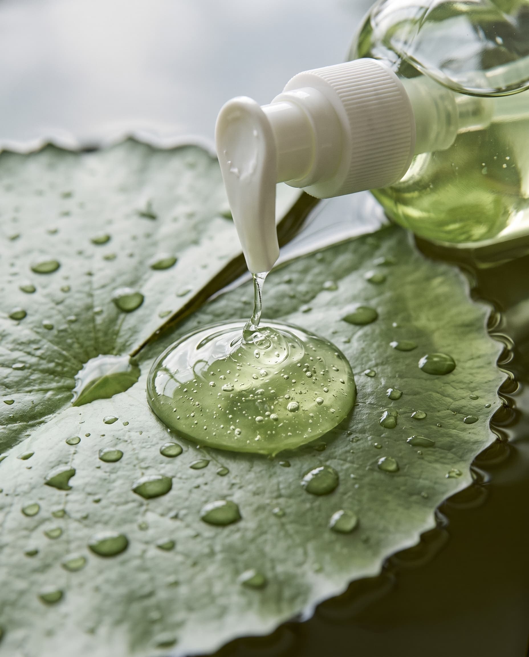 Macro of a white pump nozzle dispensing a dollop of clear green gel onto a wet lily pad, made with Nano Banana Pro