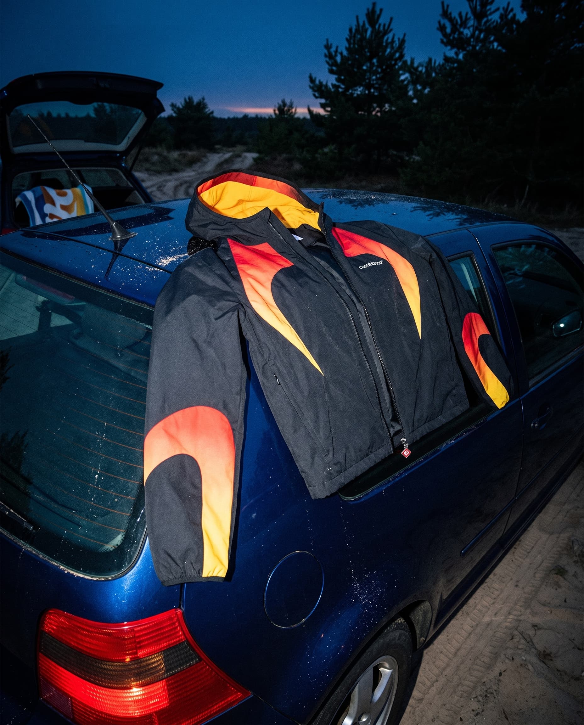 Flame-panel jacket draped over the roof of a dark blue hatchback at blue hour, lit by flash, made with Nano Banana Pro