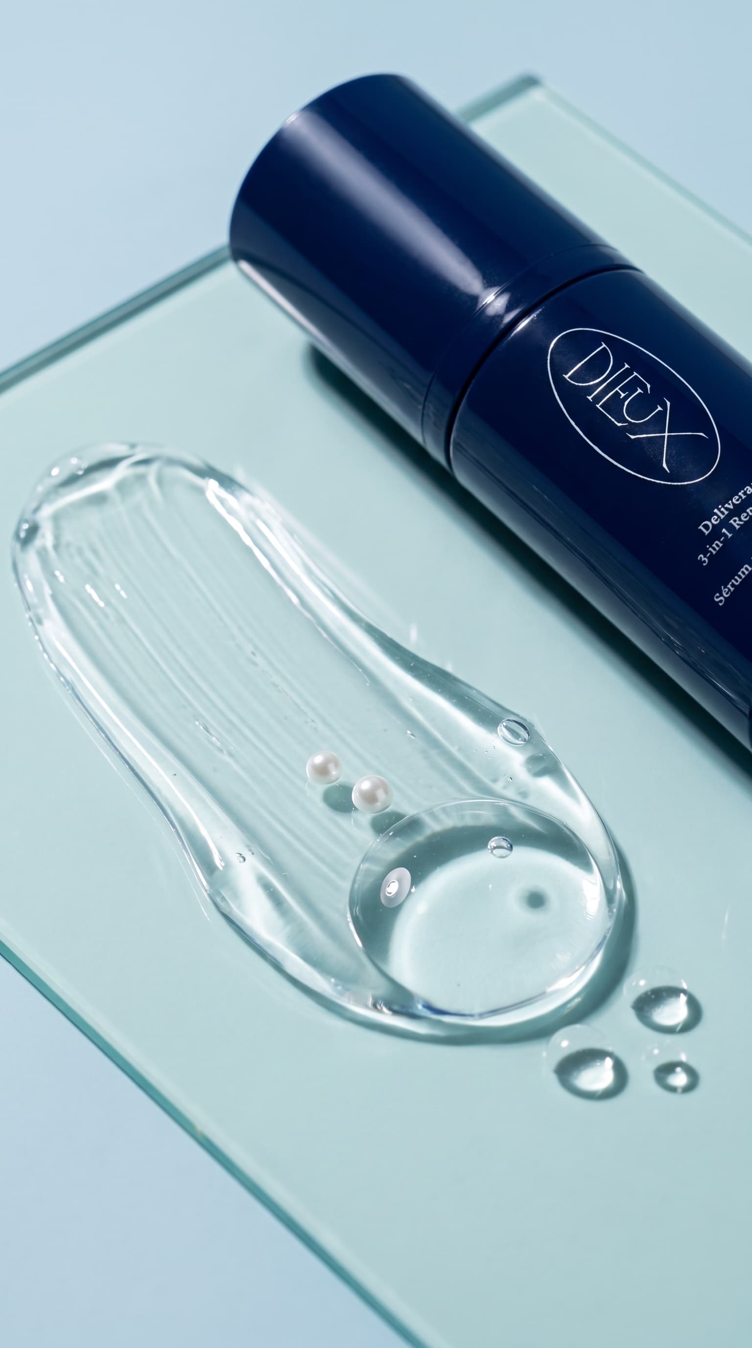 Macro of a clear serum swipe with droplets and two small pearls on pale aqua glass beside a navy bottle, made with Nano Banana Pro