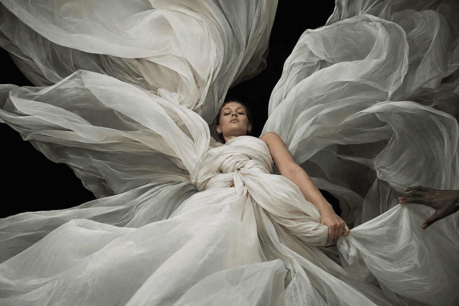 Woman wrapped in crinkled ivory organza seen from below, a second person's hand at the right edge, made with GPT Image 2.5 Sunburst