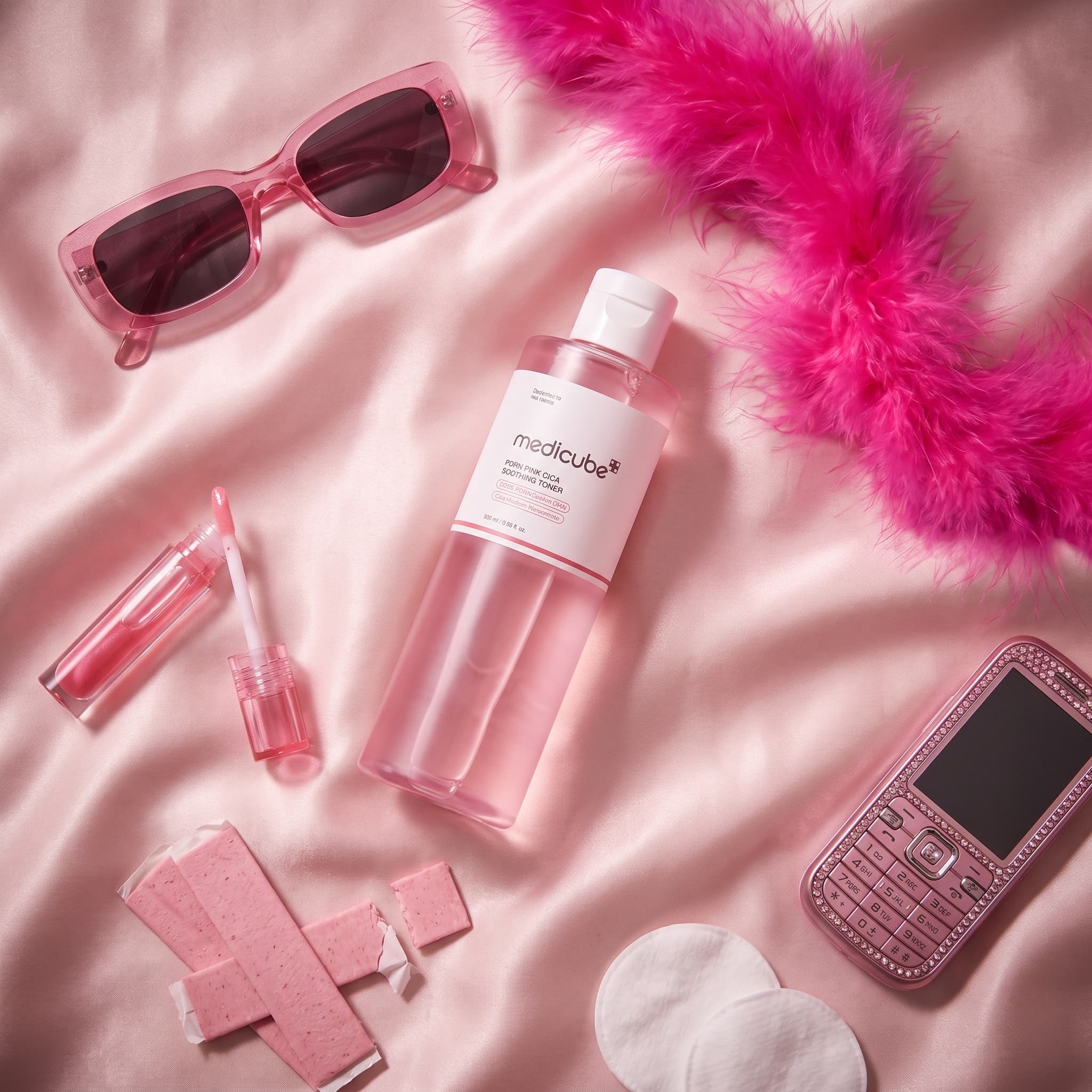 Top-down flat lay of a pink toner bottle with pink sunglasses, a feather boa, lip gloss, gum and a rhinestone phone on pink satin, made with Nano Banana Pro
