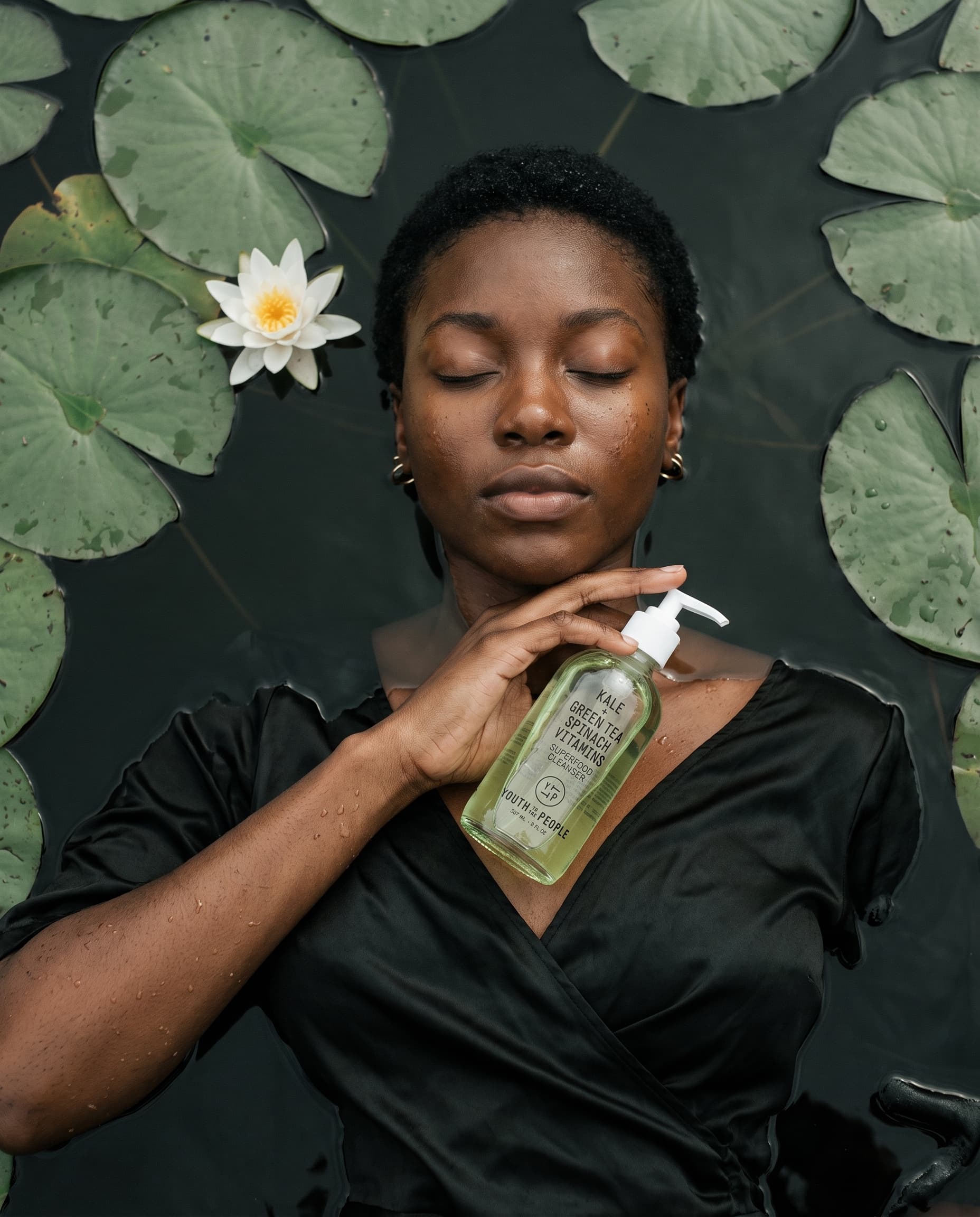 Woman floating face-up among lily pads in a black satin dress, holding a green cleanser bottle at her collarbone, made with Nano Banana Pro