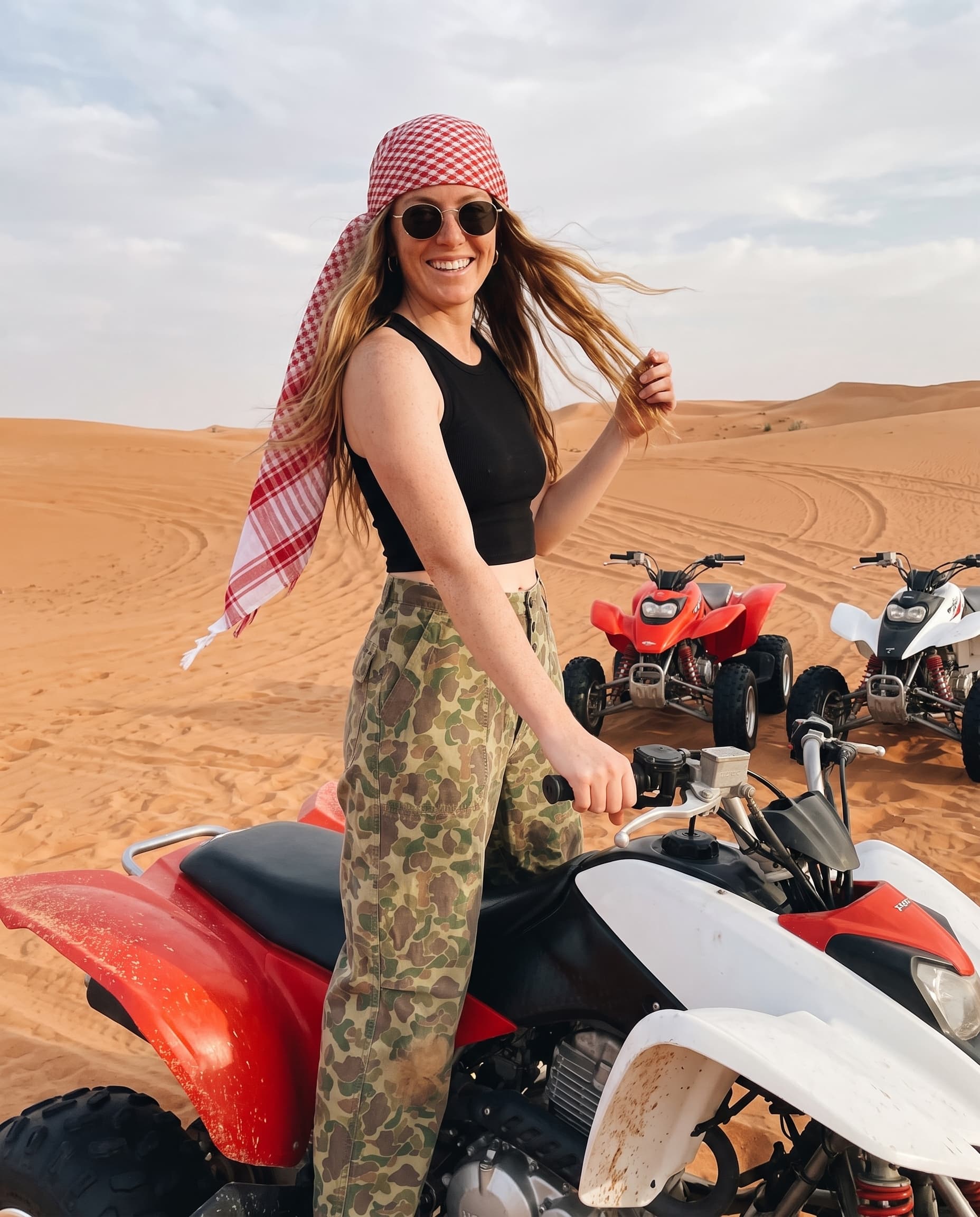 Smiling woman in camo pants and a red checked headscarf on a quad bike in the dunes, made with Nano Banana Pro