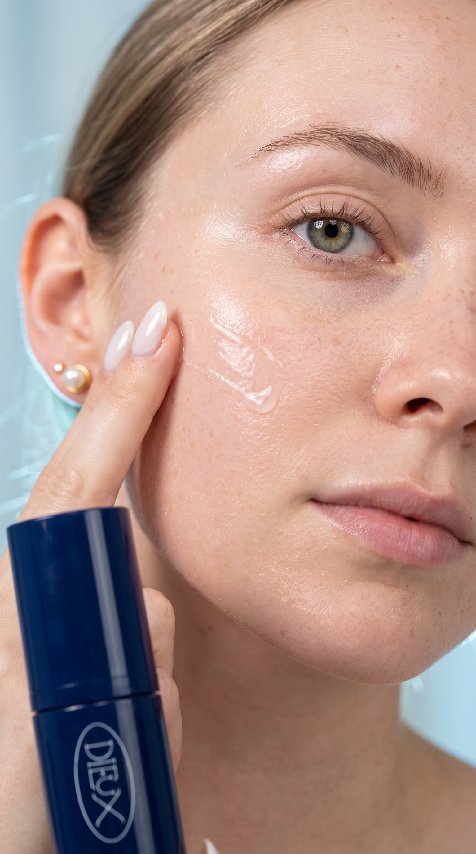 Close-up of fingertips spreading clear serum across a freckled cheekbone, navy bottle in the corner, made with Nano Banana Pro