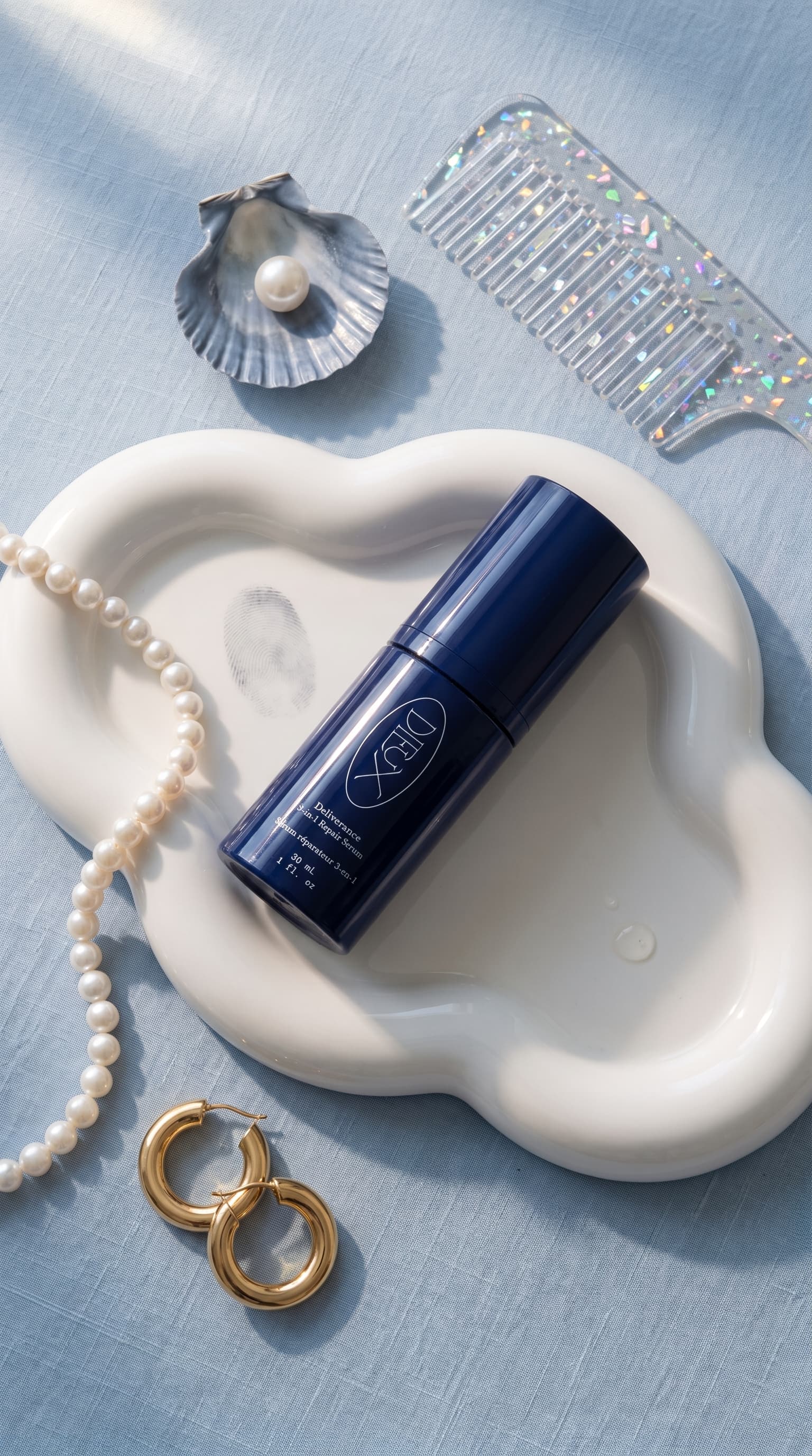 Flat lay of a navy serum bottle on a white cloud-shaped tray with pearls, gold hoops, a shell and a holographic comb, made with Nano Banana Pro
