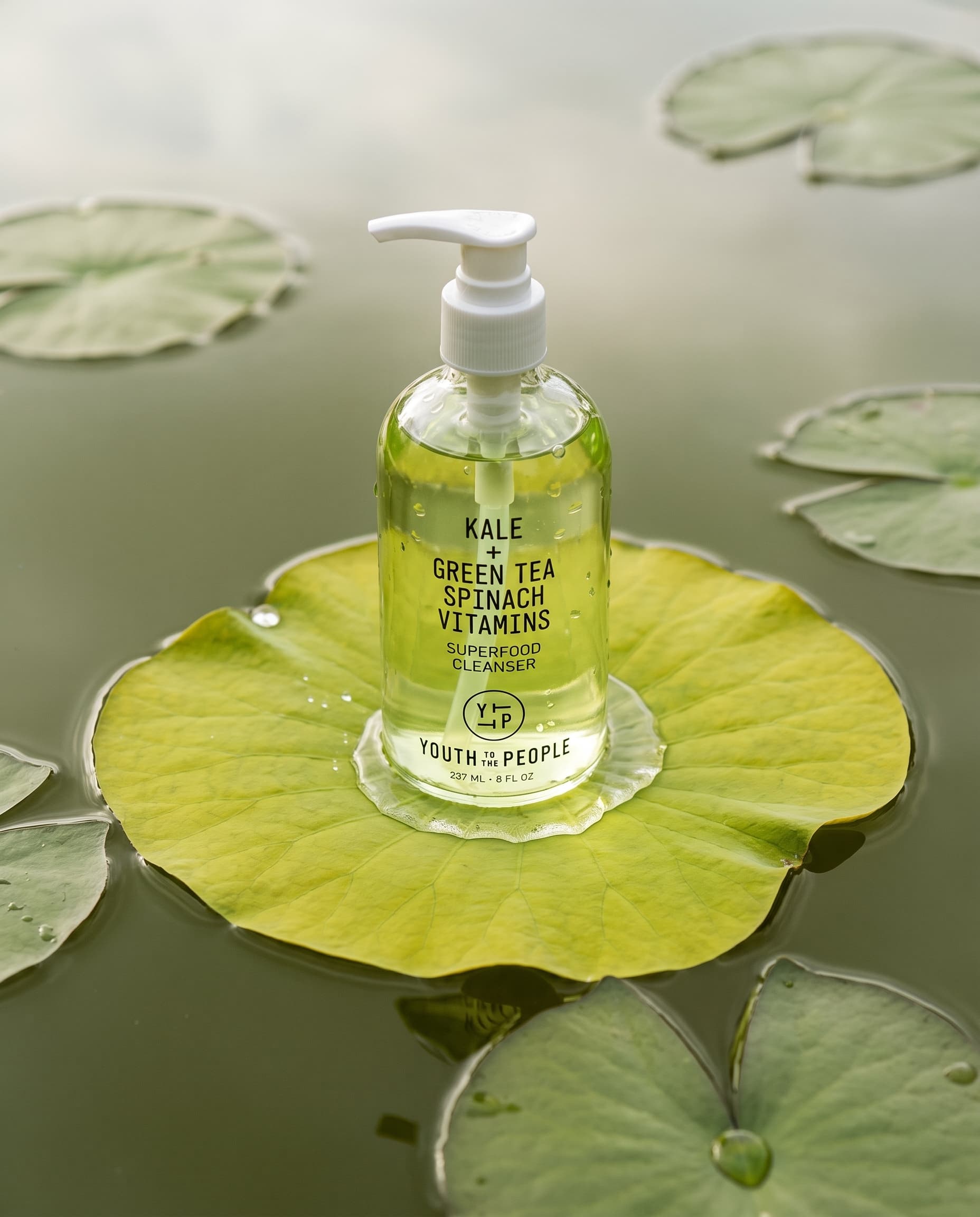 Clear glass pump bottle of green gel cleanser standing on a chartreuse lotus leaf in olive pond water, made with Nano Banana Pro