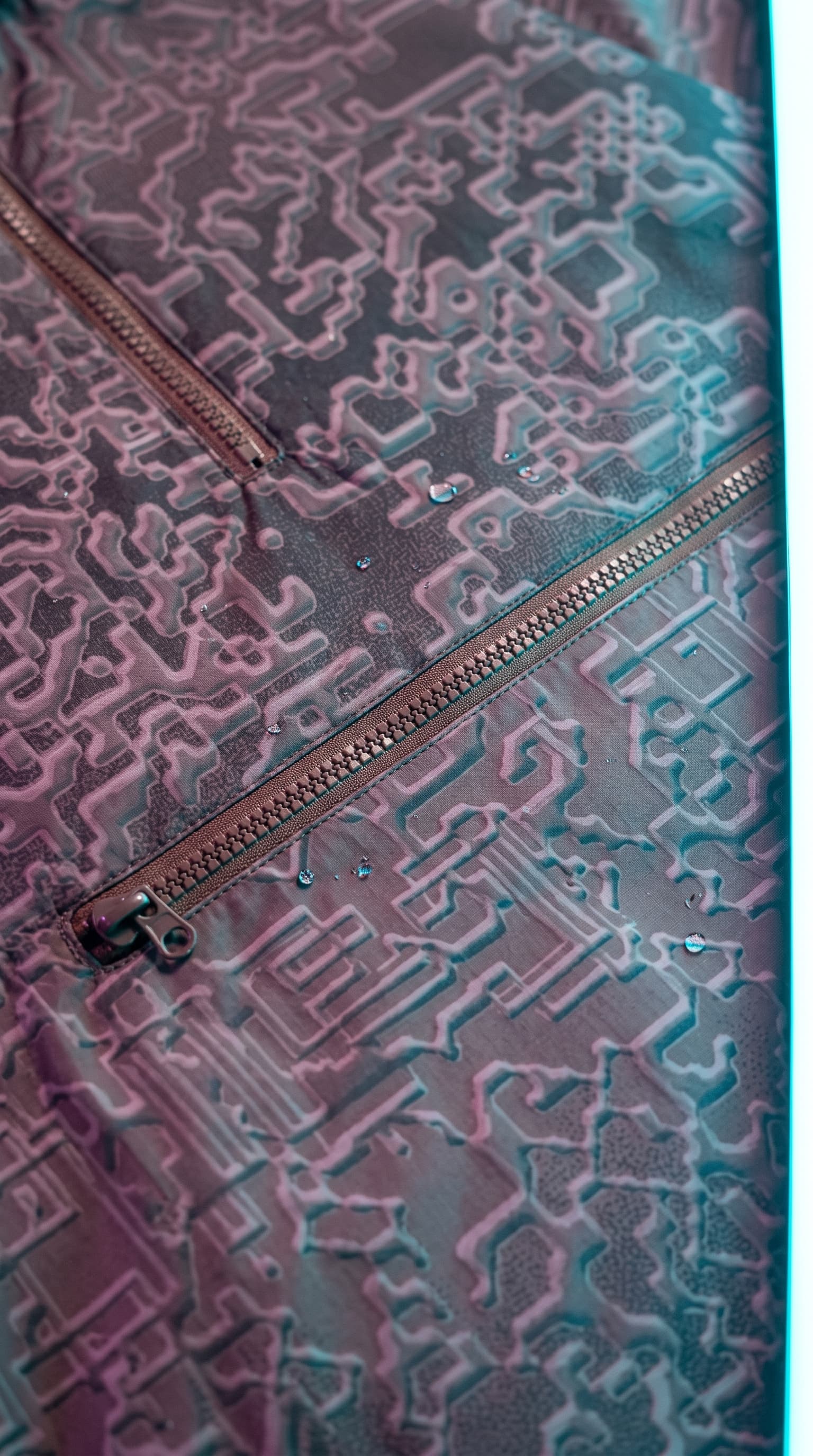 Macro of raised circuit-pattern fabric with a diagonal zip and water drops in cyan light, made with gemini