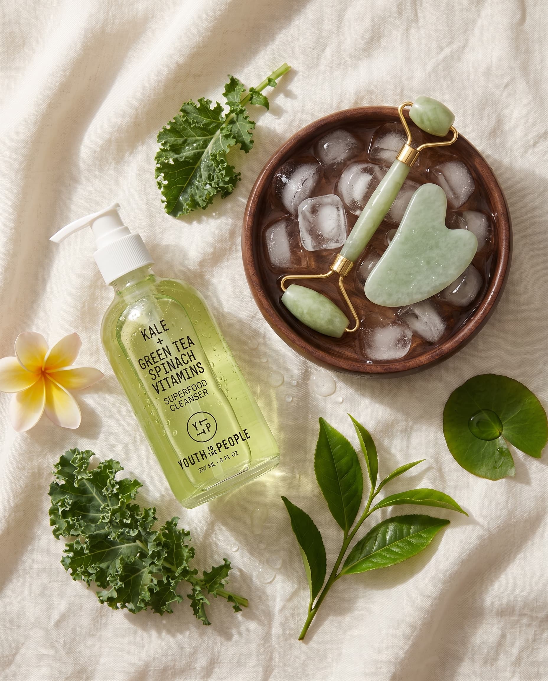Flat lay on ivory linen of a green cleanser bottle, kale, green tea leaves, a frangipani and a jade roller in a bowl of ice, made with Nano Banana Pro