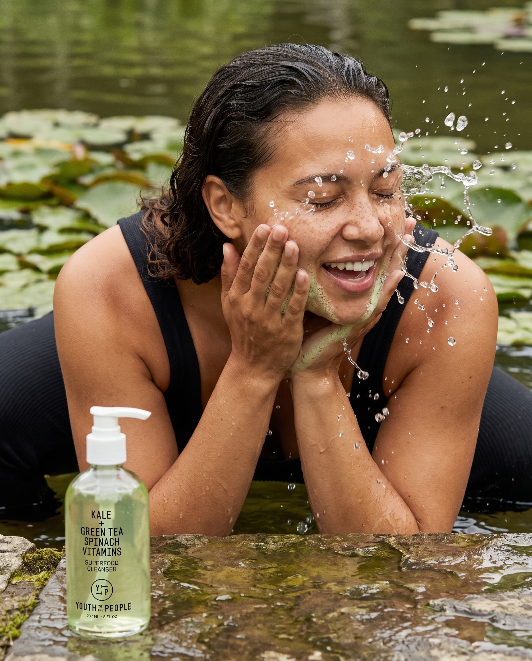 Woman laughing as she splashes pond water on her face, a green cleanser bottle on the stone edge, made with Nano Banana Pro