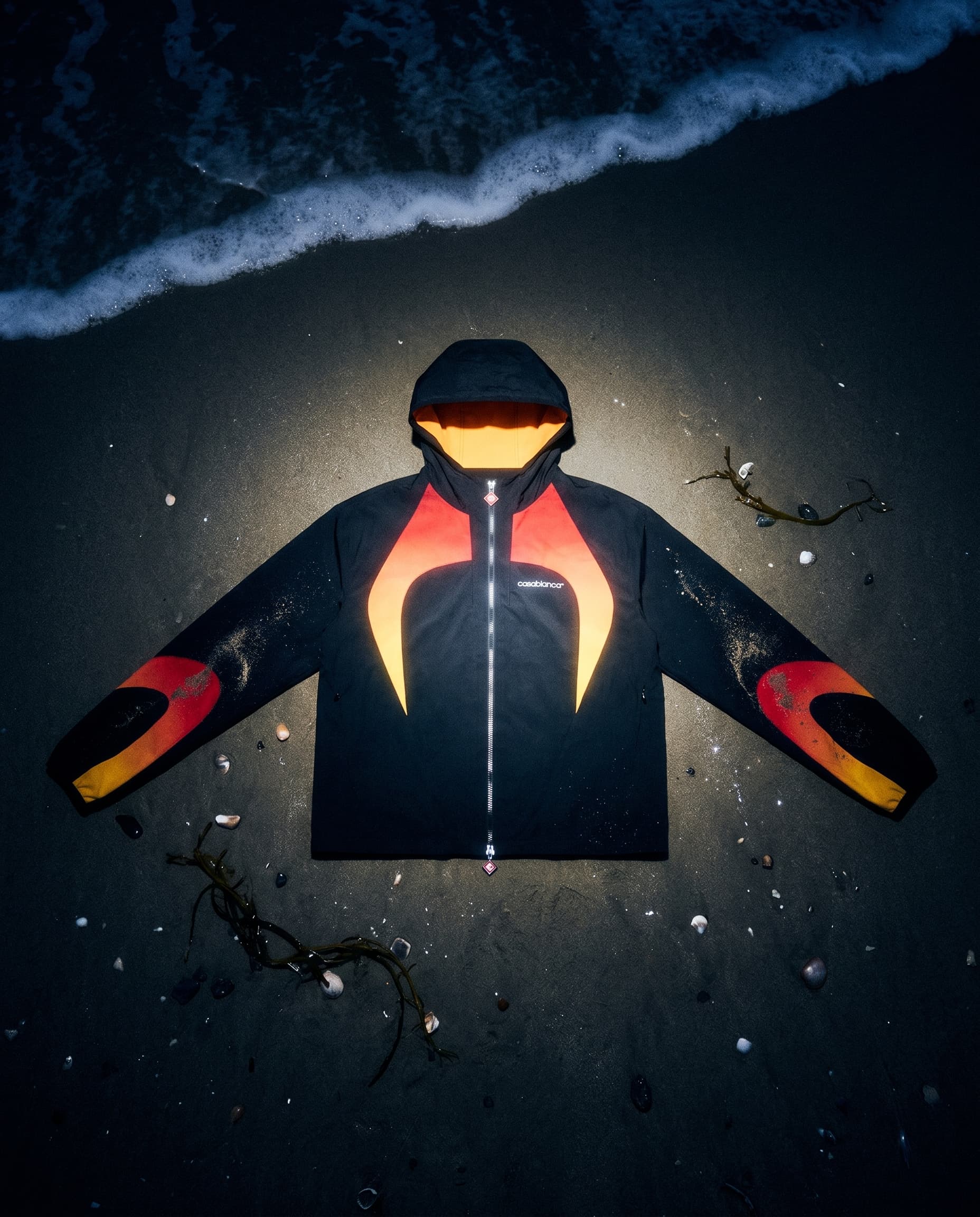 Top-down flat lay of a flame-panel jacket with arms spread on wet sand at night, sea foam at the top, made with Nano Banana Pro